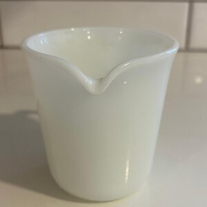 May include: A white, opaque glass creamer with a spout. The creamer has a rounded base and a flared top. The spout is small and pointed. The creamer is made of a smooth, glossy material.