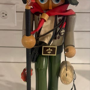 以下が含まれることがあります： 「The Boy Scout」と記された木製のくるみ割り人形の置物。金色のフルール・ド・リスのエンブレムが付いたダークグリーンの帽子、赤いネッカチーフ、ベージュの制服を着用。小さなアメリカ国旗が付いた旗竿を持ち、長方形の台座の上に立っている。
