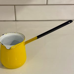 May include: A yellow enamel coffee pot with a long black handle. The pot has a white interior and a small spout. The handle is attached to the side of the pot and is angled upwards. The pot is sitting on a white surface.