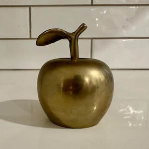 Puede incluir: Una manzana de metal de color dorado con una hoja en la parte superior. La manzana es lisa y brillante.