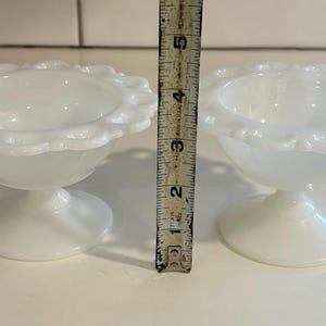 Puede incluir: Dos copas de postre blancas con pedestal y bordes con volantes. Cada copa mide aproximadamente 7,6 cm de alto. Las copas son de vidrio blanco opaco y están sobre una superficie blanca.