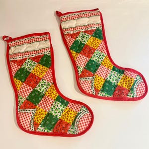 Può includere: Due calze di Natale con un design patchwork. Le calze presentano quadrati rossi, verdi e gialli, con un motivo a quadri rossi e bianchi sui bordi. La parte superiore delle calze è bianca con un motivo trapuntato e un anello rosso.