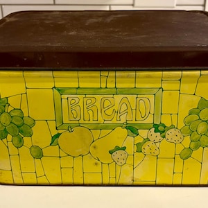 May include: A rectangular yellow bread box with a brown lid. The box is decorated with green grapes, apples, pears, and strawberries. The word "BREAD" is written in green letters on the front.