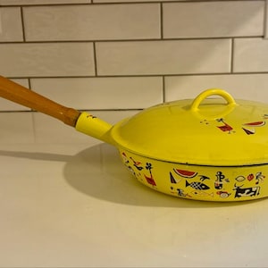 Może przedstawiać: Jasnożółta patelnia w stylu vintage z pokrywką i długim drewnianym uchwytem. Patelnia jest ozdobiona fantazyjnymi ilustracjami zwierząt, owoców i postaci. Uchwyt ma około 30 cm długości.