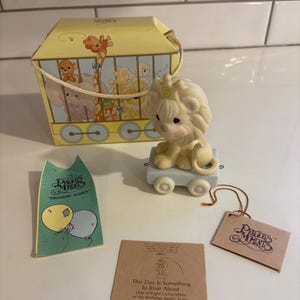 May include: A Precious Moments figurine of a lion with a number 5 on its head, sitting on a light blue cart. A yellow box with animal illustrations and the text "Birthday Series" is in the background. Includes tags.