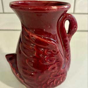 May include: A burgundy ceramic jug with a swan design. The jug features a swan's head as a handle and the body of the swan as the main body of the jug. The jug has a flared top and a small spout at the base.