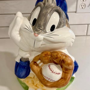 Op de afbeelding: Een keramische Bugs Bunny koekjespot in een honkbaluniform. De pot is wit met blauwe en grijze accenten, een bruine honkbalhandschoen en een honkbal. De basis is groen.
