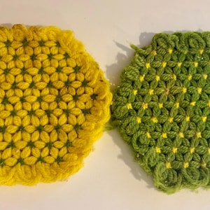 Op de afbeelding: Twee handgemaakte, zeshoekige gehaakte onderzetters. Eén is heldergeel, de andere groen. Beide hebben een bloemenpatroon en een getextureerde, geluste rand. Deze onderzetters zijn perfect om een vleugje handgemaakte charme toe te voegen.