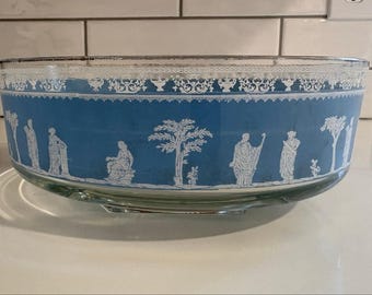 Può includere: Una ciotola rotonda in vetro con un'ampia fascia blu all'esterno. La fascia blu è decorata con figure in rilievo bianco di persone e alberi in stile classico. Un motivo decorativo bianco corre lungo il bordo superiore della ciotola.