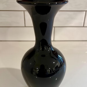 May include: A glossy black vase with a flared top and a rounded body. The vase has a smooth, reflective surface, and it sits on a white surface. The background features white tiles.