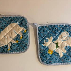 Peut inclure: Deux maniques carrées matelassées aux coins arrondis avec un liseré gris clair. Le tissu présente un fond bleu avec un petit motif floral blanc. Une manique montre un cochon blanc, tandis que l'autre affiche une vache blanche avec une fleur jaune.