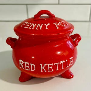 May include: A bright red ceramic kettle-shaped container with a lid. The lid is labeled "PENNY POT" in white lettering, and the body reads "RED KETTLE". The container has a handle on the lid and two side handles.