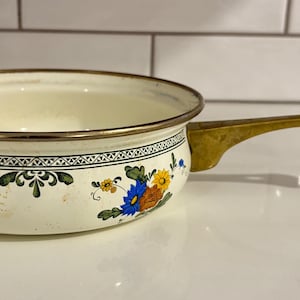 Op de afbeelding: Crèmekleurige emaille steelpan met een messing handvat en rand. De pan heeft een bloemenontwerp met blauwe, gele en oranje bloemen. De handgreep is ongeveer 20 cm lang. Een vintage keukengerei.