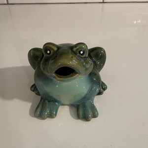 Könnte beinhalten: Eine Keramikfrosch-Figur mit glänzender Oberfläche, die einen dunkel olivgrünen Kopf und Rücken aufweist, der in eine hellere, blass blaugrüne Brust und einen Bauch übergeht. Der Frosch hat ein weit geöffnetes Maul und vier Schwimmfüße.