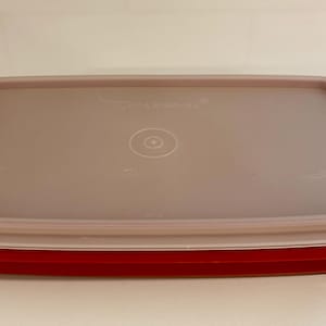 May include: A rectangular food storage container with a red base and a translucent lid. The lid has a light brown tint and a circular design in the center. The container is designed for food storage.