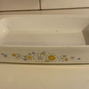Puede incluir: Fuente rectangular blanca para hornear con un diseño floral en amarillo, azul y blanco. El plato tiene un borde ligeramente elevado y probablemente está hecho de cerámica o vidrio. Un artículo de cocina.