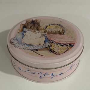 Può includere: Una scatola rotonda rosa con un coperchio con un'illustrazione di topi. Il coperchio ha un bordo rosa chiaro e una scena di topi in un cesto. Il lato della scatola è decorato con un motivo floreale blu. La scatola è di metallo.