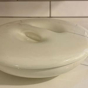 May include: A white, oval-shaped decorative dish with a central indentation. The dish is approximately 5 inches in diameter and has a smooth, glossy finish. A measuring tape is visible in the background.