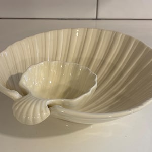May include: Cream-colored ceramic serving dish set. The set includes a large, shell-shaped dish with a smaller, shell-shaped dip bowl attached. The dish has a ribbed texture, and the set is designed for serving appetizers or snacks.