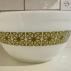 Puede incluir: Bol para mezclar blanco con una banda decorativa de motivos florales verde oliva. El bol tiene forma redondeada y está hecho de un material grueso y opaco. Un artículo de cocina clásico.