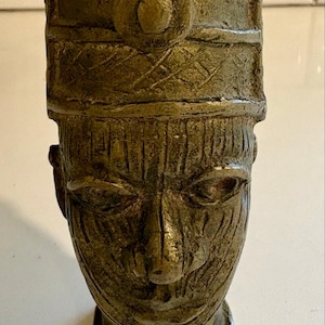 Puede incluir: Una escultura de color bronce de una cabeza humana con un tocado cilíndrico. La cara tiene rasgos detallados, incluyendo ojos, nariz y boca. El tocado tiene un diseño circular en la parte delantera y un patrón texturizado.