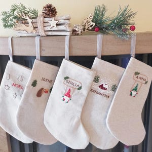 Op de afbeelding: Vijf gepersonaliseerde kerstsokken met geborduurde namen en ontwerpen. De sokken zijn beige met witte borduursels. De namen op de sokken zijn Noel, Irene, Giulia, Samantha en Anna.