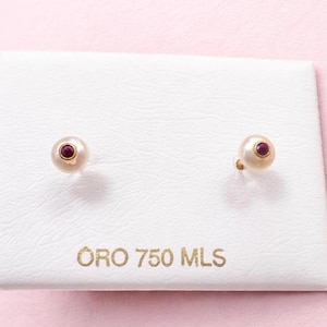 Peut inclure: Boucles d'oreilles à clous dorées avec un aspect perlé, chacune ornée d'une petite pierre précieuse rouge et ronde. Les boucles d'oreilles sont présentées sur une surface blanche avec le texte "ORO 750 MLS" en lettres dorées, sur fond rose.