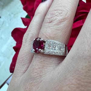 May include: A ring with a large, round, red ruby gemstone surrounded by a band of smaller, white diamonds.