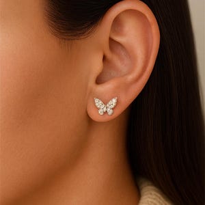 Può includere: Primo piano di un orecchino a forma di farfalla. L'orecchino è composto da piccole pietre scintillanti, probabilmente diamanti o zirconi, disposte in un design delicato e simmetrico. L'orecchino è su un orecchio.