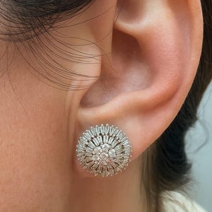 May include: A close-up of a dazzling, circular stud earring. The earring showcases a central cluster of small, round gems encircled by a halo of baguette-cut stones, producing a sunburst design. The earring is set against the backdrop of a person's ear.