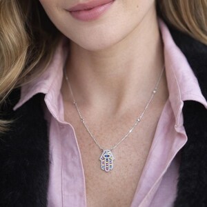 May include: A silver necklace featuring a Hamsa hand pendant. The pendant is adorned with colorful gemstones, including blue, pink, yellow, and white. The necklace is worn against a pink collared shirt and a black jacket.