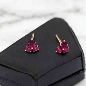 Può includere: Un paio di orecchini d'oro con tre pietre preziose rosa ciascuno, esposti su una scatola di gioielli in velluto nero.