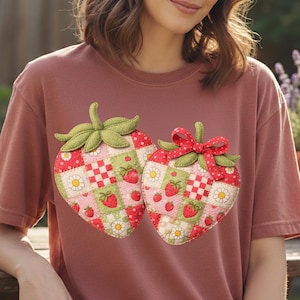 Könnte beinhalten: Staubrosafarbenes T-Shirt mit zwei aufgenähten Erdbeeren. Die Erdbeeren bestehen aus Patchwork-Quadraten in Rot, Grün und Weiß, mit Gänseblümchen- und Erdbeerdetails. Eine Erdbeere hat eine rote Schleife.