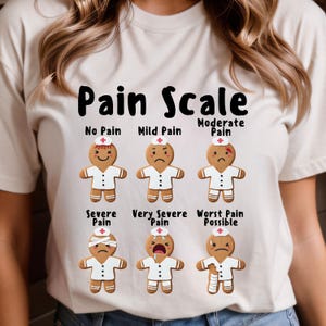 Könnte beinhalten: Cremefarbenes T-Shirt mit der Aufschrift "Pain Scale" und Lebkuchenmännchen, die Schmerzstufen von "No Pain" bis "Worst Pain Possible" darstellen. Jedes Lebkuchenmännchen trägt eine Krankenschwestermütze.