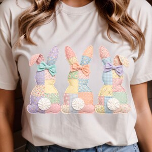Könnte beinhalten: Cremefarbenes T-Shirt mit drei aufgenähten Hasen. Jeder Hase besteht aus Patchwork-Stoff in Pastellfarben, mit einer Schleife am Hals und einem flauschigen weißen Schwanz. Die Hasen sind in einer Reihe auf der Vorderseite des Shirts angeordnet.