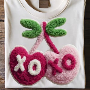 May include: Cream-colored t-shirt featuring a textured, embroidered design of cherries and a dragonfly. The cherries are pink and fuchsia with white and pink "XOXO" lettering. The dragonfly has green wings and a pink body.