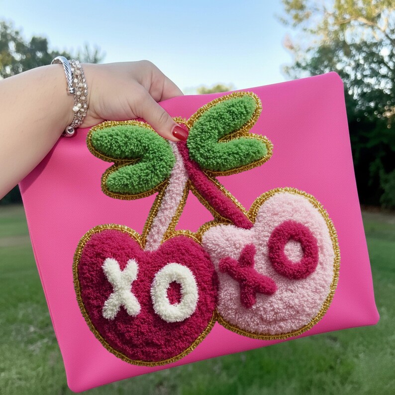 May include: A bright pink clutch featuring a cherry design with the letters "XO". The cherries are embroidered with a textured, plush material in shades of pink and red, with green leaves and gold trim. The clutch is held up against a background of green grass and a blue sky.