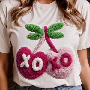 May include: White t-shirt featuring a whimsical design of two heart-shaped cherries with the letters "XO" in white. The cherries are in shades of pink and the stem and leaves are green. The design has a textured, plush appearance.