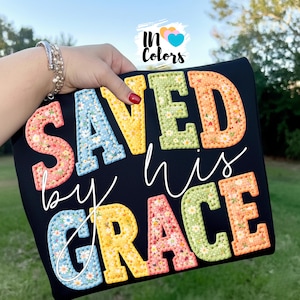 Puede incluir: Camiseta negra con la frase "SAVED by his GRACE" en letras coloridas con estampado floral. El diseño incluye el texto "In Colors" con un logotipo de corazón. La camiseta es sostenida por una persona.