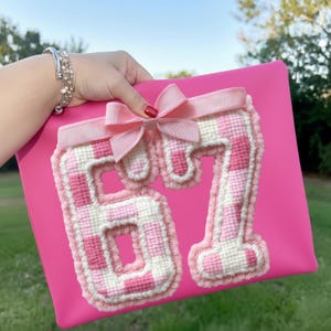 以下が含まれることがあります： ピンク色のクラッチバッグ。ピンクと白のギンガムチェック模様で刺繍された数字の「67」が特徴。数字は白いトリムで縁取られ、上部にはピンクのリボンが付いています。
