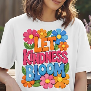 Puede incluir: Camiseta blanca con la frase "LET KINDNESS BLOOM" en letras coloridas y cosidas. El diseño incluye flores vibrantes en rosa, naranja y azul con hojas verdes, creando un ambiente alegre y positivo.