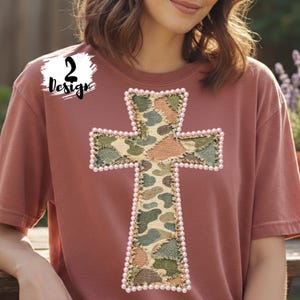 May include: A dusty rose-colored t-shirt featuring a large cross design. The cross is made of camouflage fabric with a pearl-like trim. The upper left corner of the shirt has the text "2 Design" in a white box.