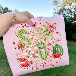 Könnte beinhalten: Rosa Sweatshirt mit dem in Rot gestickten Schriftzug "Sweet Summer Time". Die Buchstaben sind mit Wassermelonenscheiben und grünen Vichymustern verziert. Das Design umfasst Wassermelonenscheiben, Schleifen und Herzen.