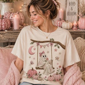 Może przedstawiać: Kremowy t-shirt z krótkim rękawem z nadrukiem graficznym przedstawiającym ducha w różowej kokardce, siedzącego na huśtawce zawieszonej na gałęzi drzewa. Wzór zawiera różowe dynie, gwiazdy, półksiężyc, motyle i wiszące girlandy.