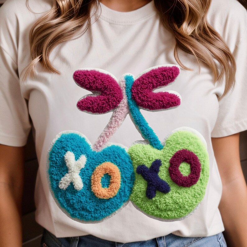 May include: White t-shirt featuring a colorful, textured cherry design. The cherries are blue and green, with the letters "XOXO" in white, orange, and purple. The design also includes a dragonfly with pink and purple wings.