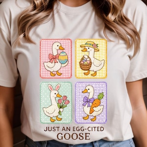 May include: A cream-colored t-shirt featuring four cartoon geese in colorful squares. Each goose is decorated with Easter-themed accessories, including eggs, a basket, bunny ears, and carrots. The text "JUST AN EGG-CITED GOOSE" is printed below the images.
