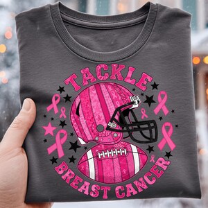 以下が含まれることがあります： ピンク色のフットボールヘルメットとボール、ピンクのリボンと星が描かれた濃い灰色のスウェットシャツ。「TACKLE BREAST CANCER」の文字がピンク色でデザインの周りにプリントされています。デザインはシャツの前面の中央に配置されています。