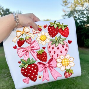 Può includere: Una federa azzurra con ricami di fragole, fiocchi e margherite. Le fragole sono rosse e rosa con foglie verdi. I fiocchi sono rosa e gialli con motivi a quadretti e pois. Piccoli cuori rossi sono sparsi.