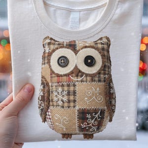 May include: Cream-colored t-shirt featuring a patchwork owl design. The owl has large button eyes and is made from various fabrics, including plaid and burlap. The design includes snowflake and swirl details.