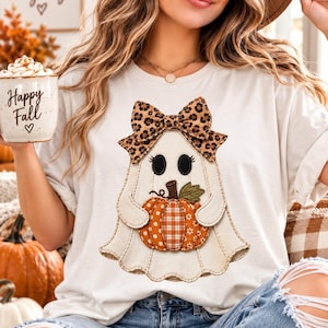Op de afbeelding: Een wit t-shirt met een afbeelding van een geest met een strik met luipaardprint, die een oranje pompoen met ruitjespatroon vasthoudt. De geest heeft zwarte ovale ogen en gestikte randen. Het ontwerp is gecentreerd op de borst van het t-shirt.